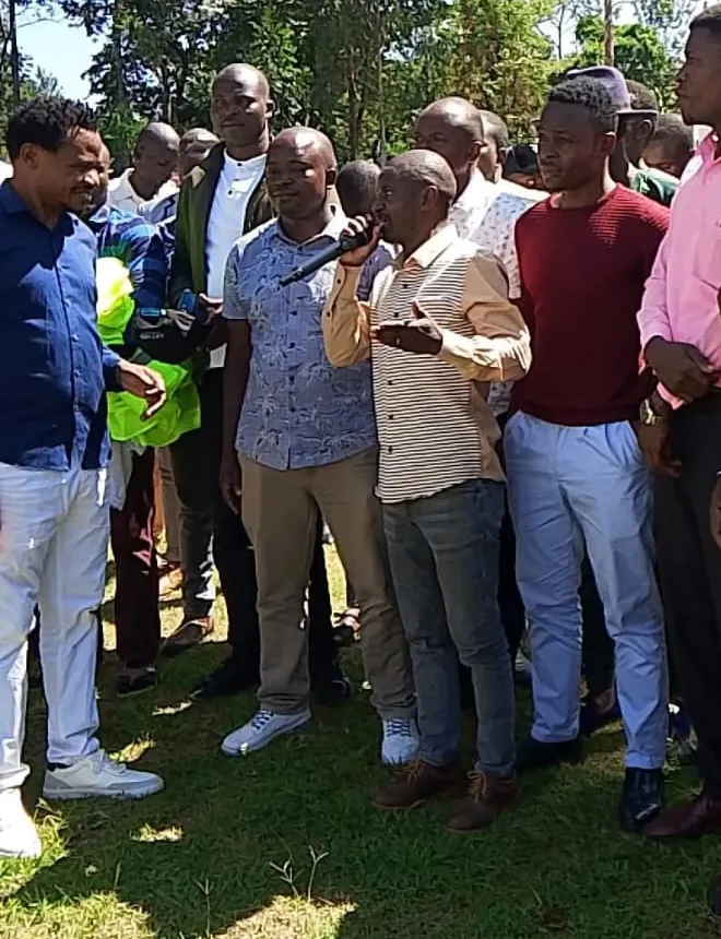Residents gathered outdoors under trees, listening to a speaker holding a microphone